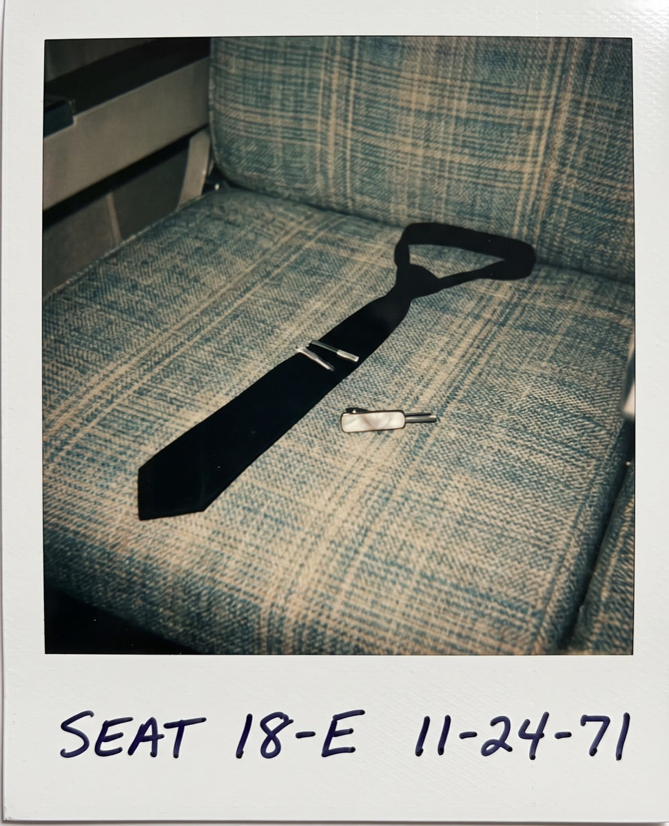 Polaroid of an empty airliner row with a dark clip-on tie left on the seat, captioned seat 18E.